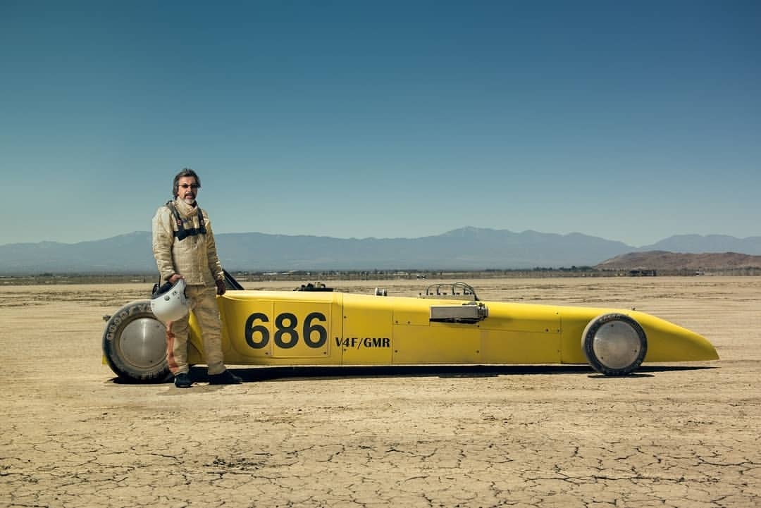 Man in racing suit stands with a yellow land speed record car on cracked desert, with mountains and blue sky in the distance.